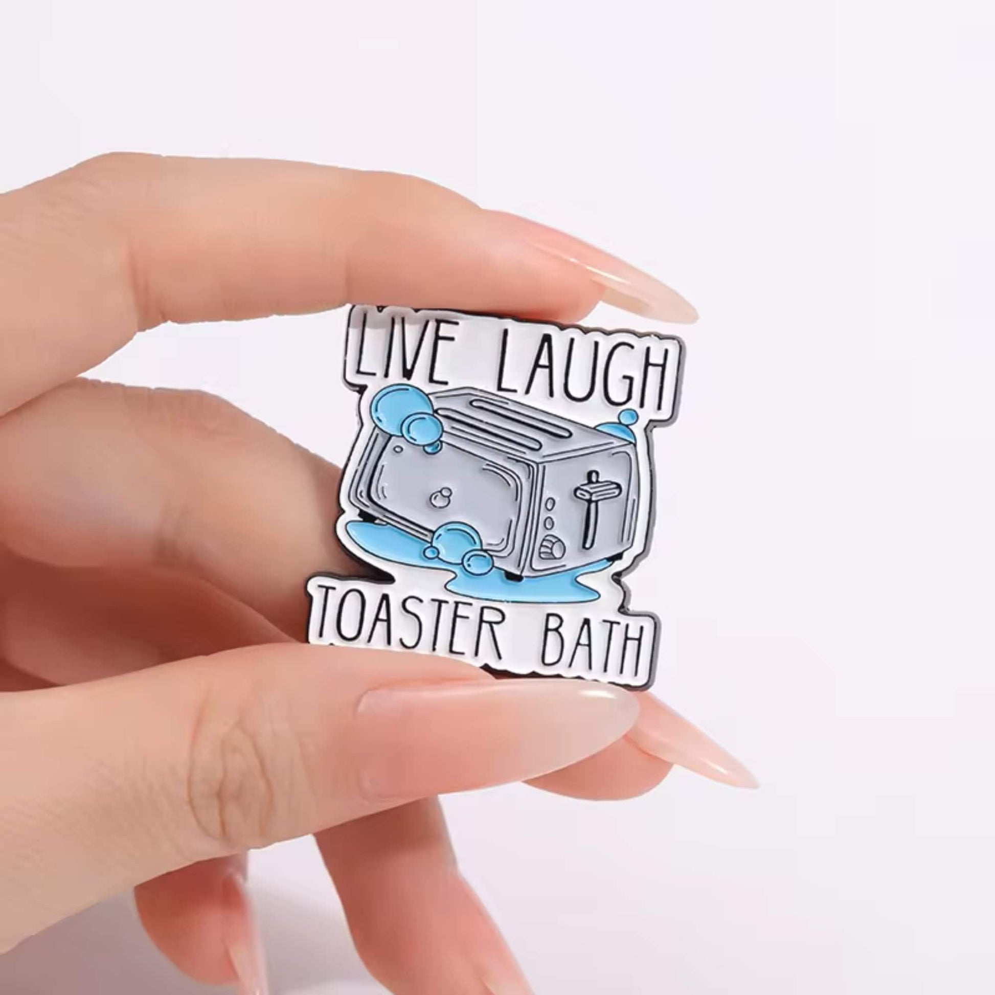A hand holding a colorful enamel pin with the text 'LIVE LAUGH TOASTER BATH' on it.