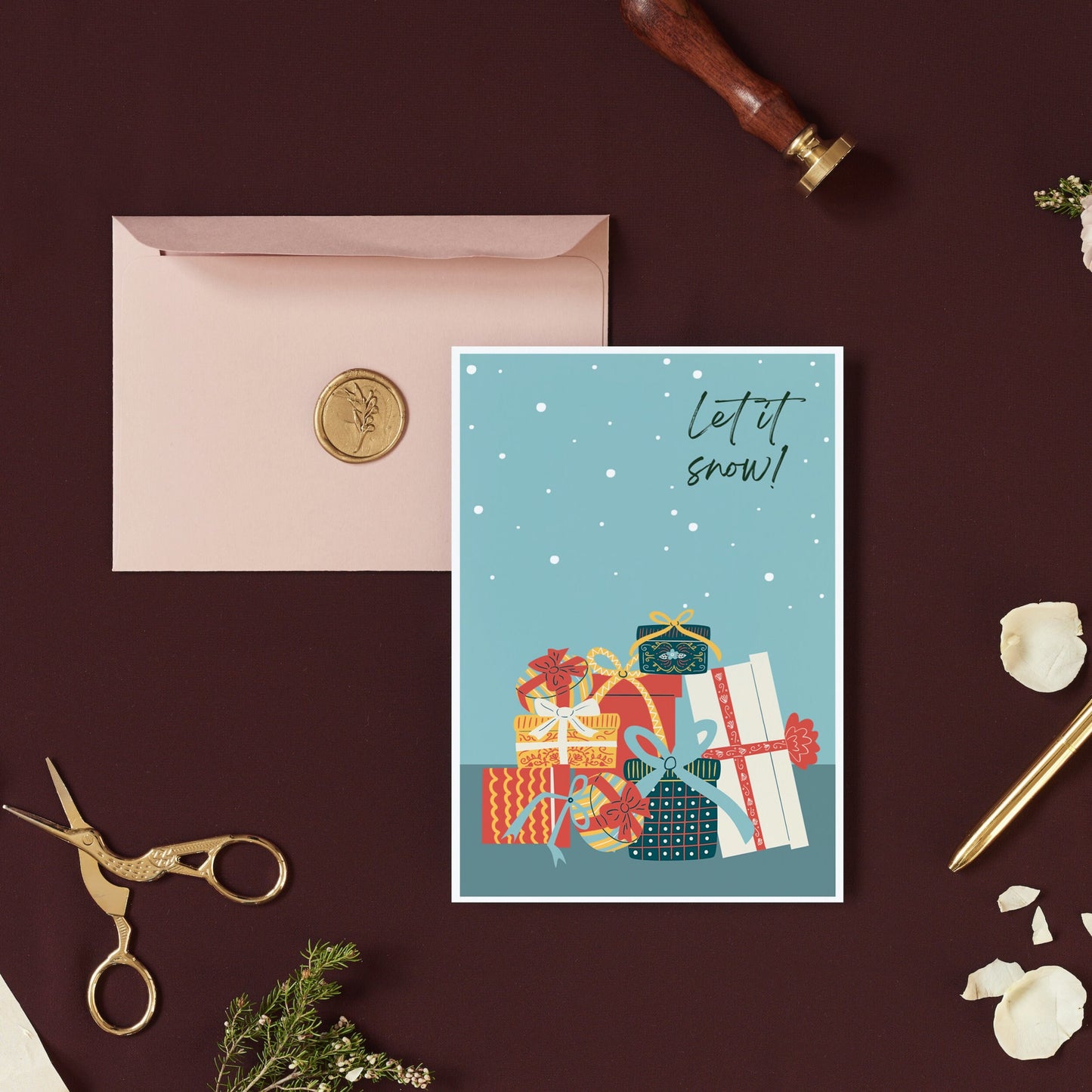 A Christmas card with a simple design featuring snowflakes and presents, placed on a desk with a pink envelope and a pair of scissors.