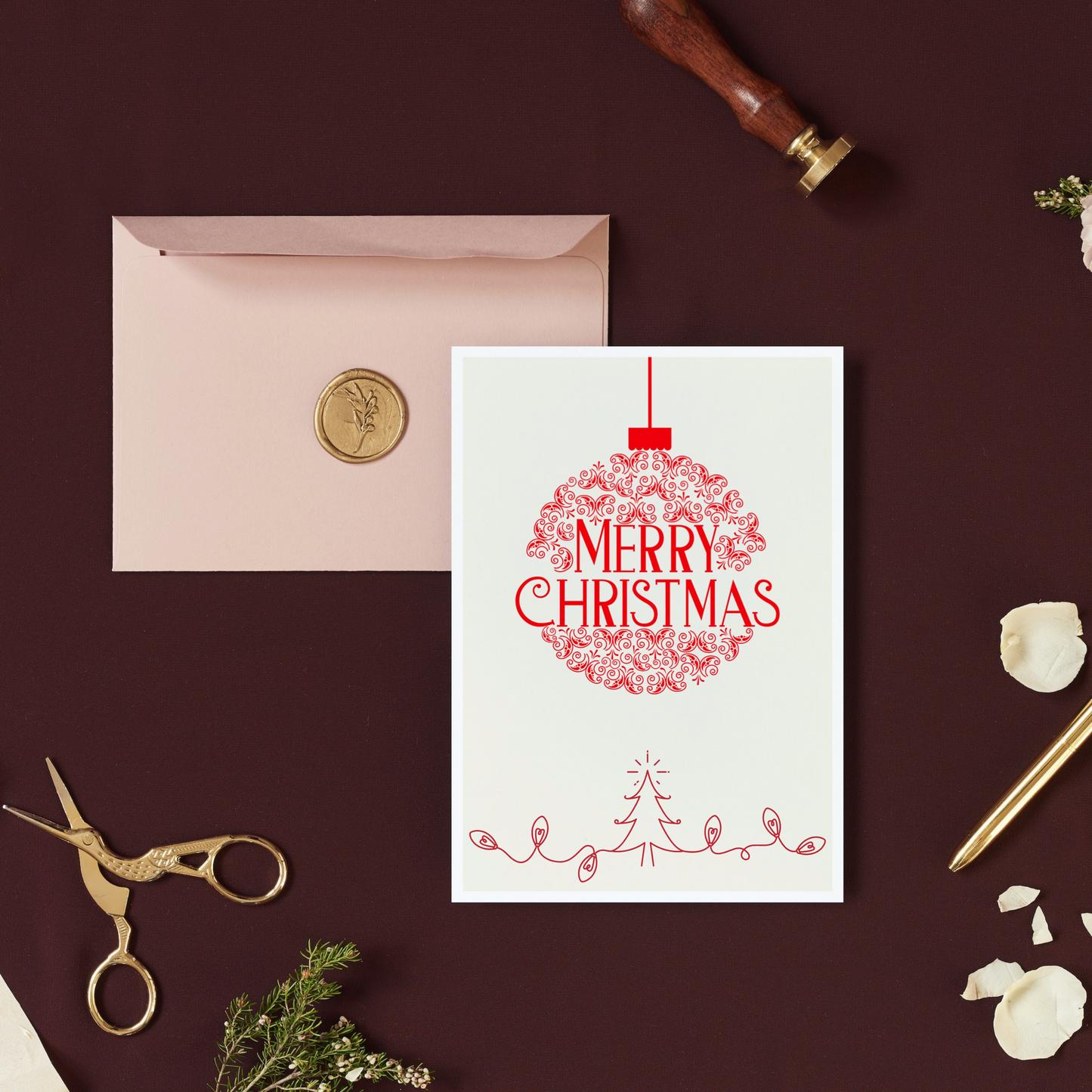 A simple red and white Christmas card with the text 'Merry Christmas' and festive decorations on the front, placed on a desk with a pen, scissors, and a cardboard cutout Christmas tree.