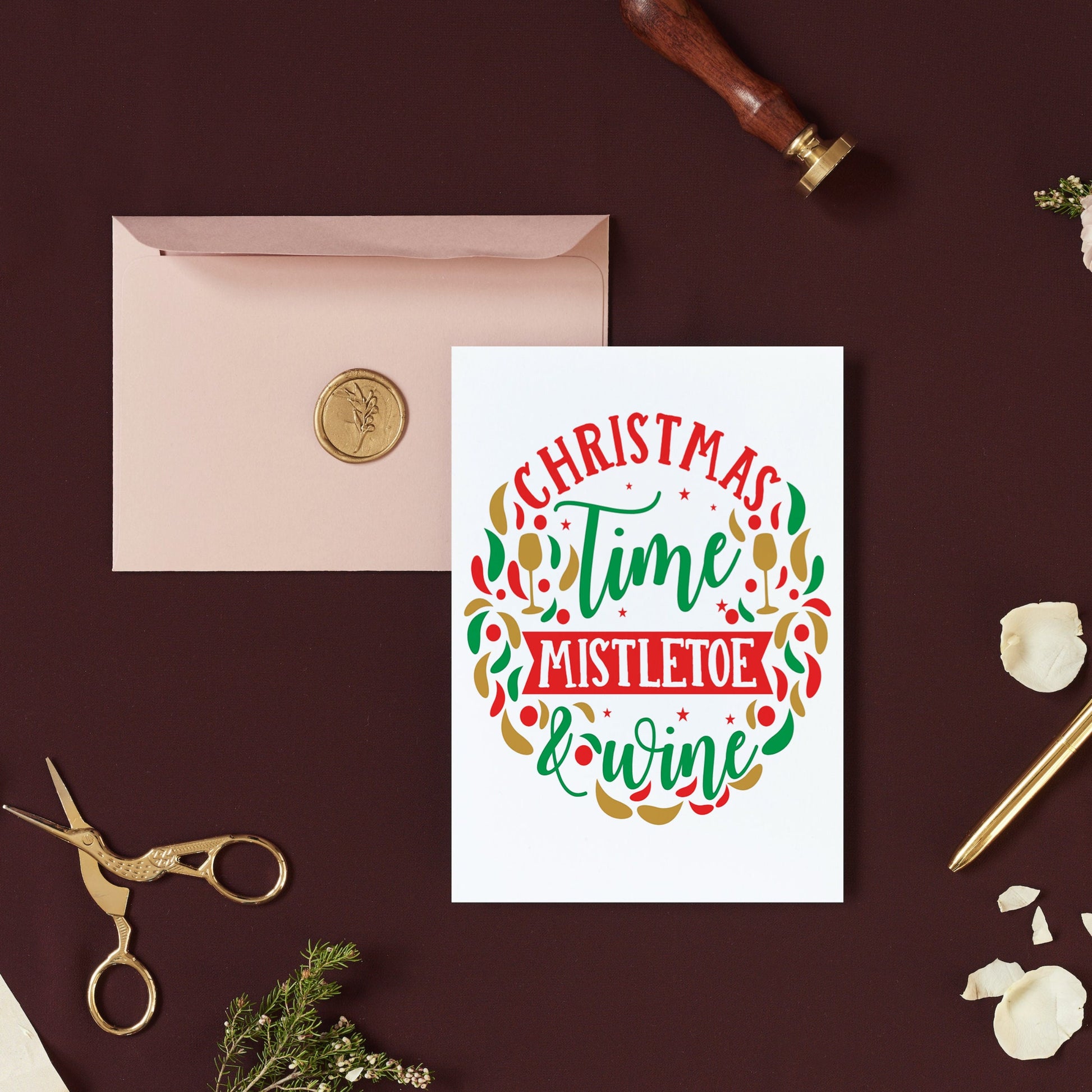 A Christmas card with 'Mistletoe and Wine' text, decorated with a Christmas tree and gifts, placed on a table with a pink envelope and a gold-colored pen.