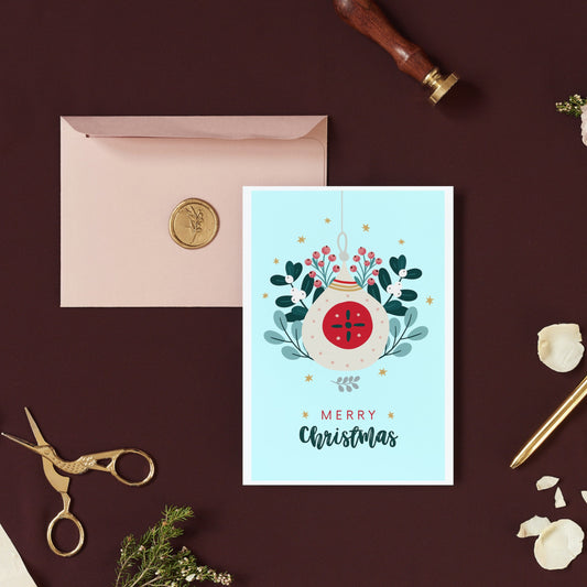 A simple Christmas card with a festive design, placed on a desk with a pen, scissors, and a pink envelope in the background.