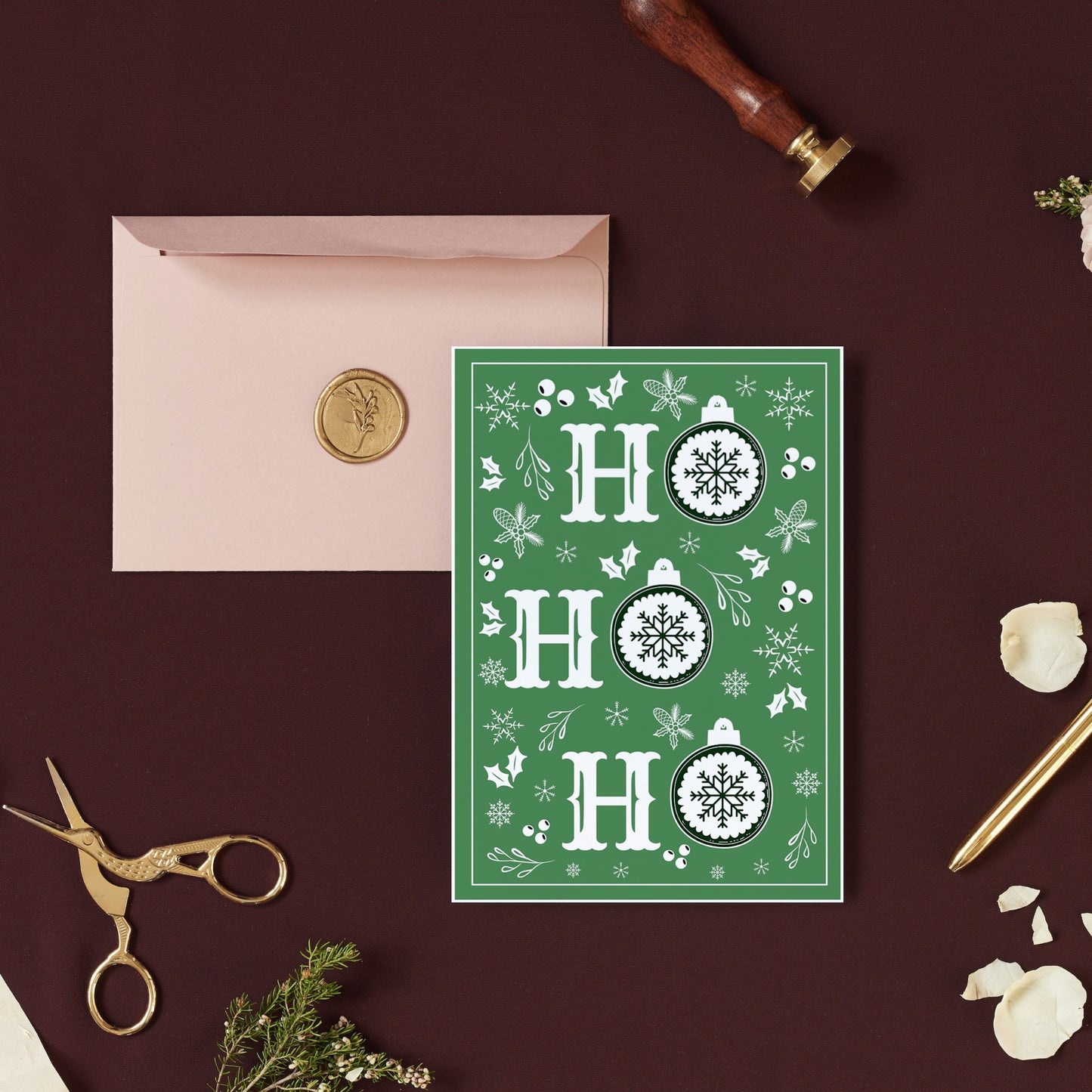 A simple Christmas card with a green background, white snowflake patterns, and the text 'ho ho' in a circular design. The card is placed on a table with a pink envelope, a pen, and a golden object for scale.