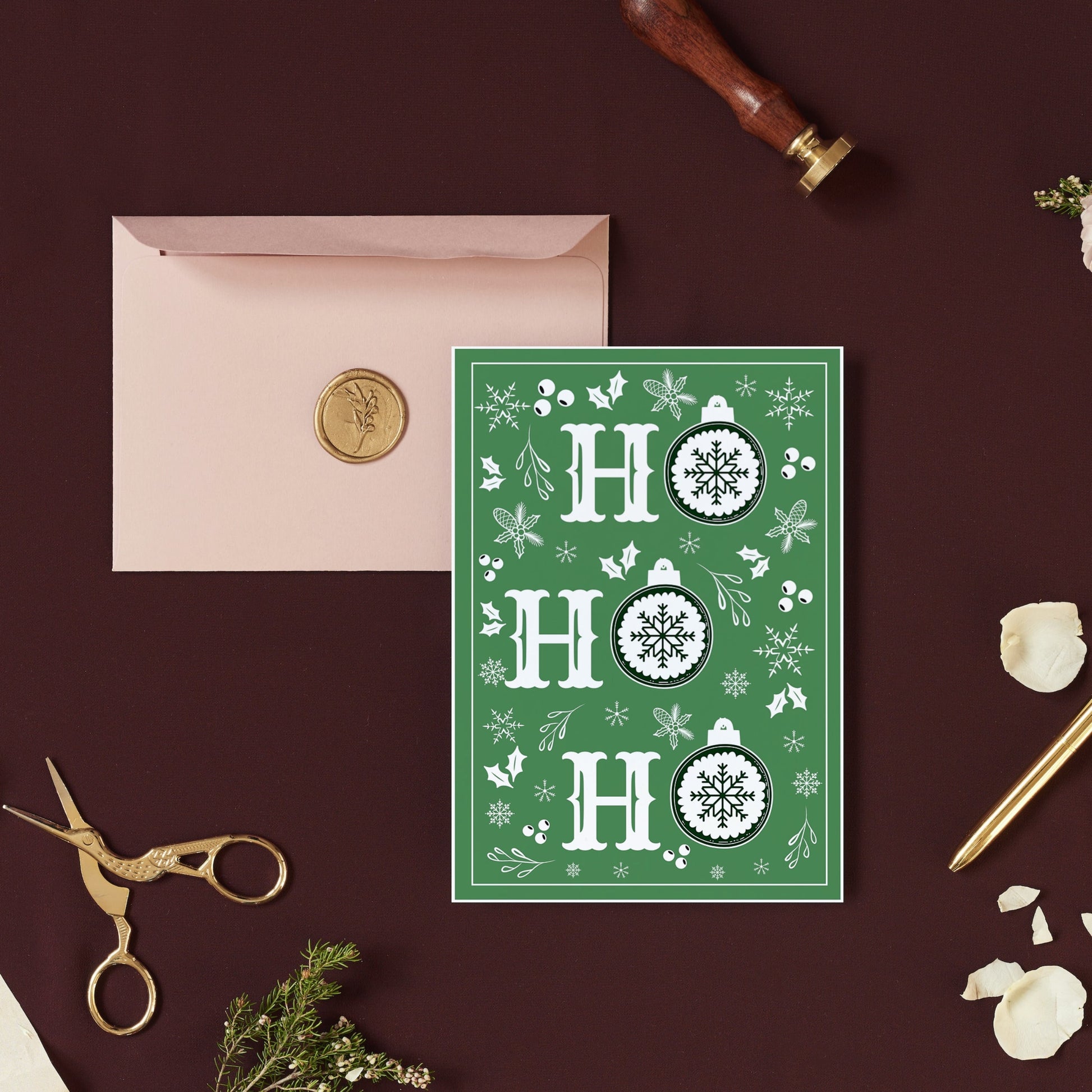 A simple Christmas card with a green background, white snowflake patterns, and the text 'ho ho' in a circular design. The card is placed on a table with a pink envelope, a pen, and a golden object for scale.