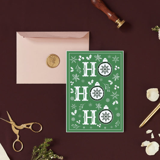 A simple Christmas card with a green background, white snowflake patterns, and the text 'ho ho' in a circular design. The card is placed on a table with a pink envelope, a pen, and a golden object for scale.